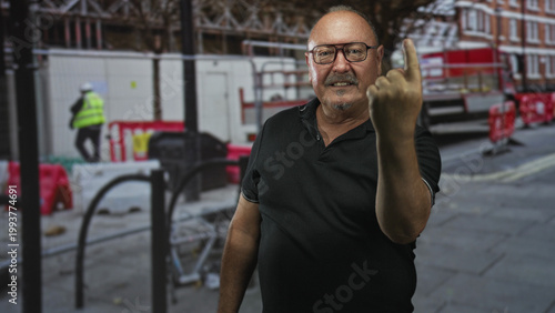 Man senior hispanic showing raised middle finger beside construction barriers and scaffolding on a street; defiance confrontation.