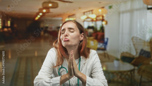 Bild auf Leinwand Woman doctor in white coat with stethoscope presses hands together in prayer inside a hotel lobby building; pleading hope