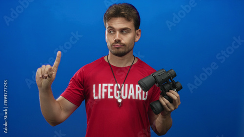 Фотография Lifeguard man wearing lifeguard shirt and whistle, points finger while holding binoculars in blue studio; vigilant duty focus