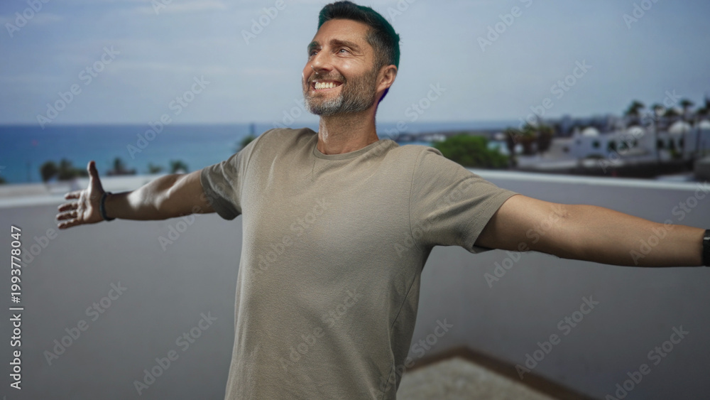 Fototapeta premium Man with arms outstretched showing hands on building rooftop terrace overlooking sea, smiling and looking up; joy escape calm.