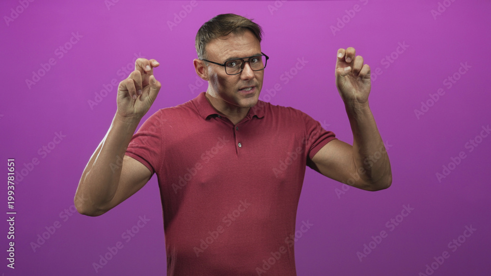 Fototapeta premium Man wearing glasses and red polo showing crossed fingers with both hands, hands raised in a purple studio setting; hopeful luck.