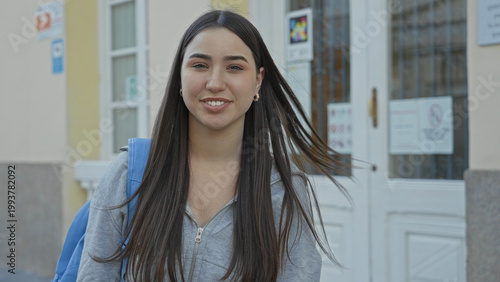 Foto Young hispanic woman smiles with a blue backpack at a building entrance, long hair visible and hands relaxed; optimism student life