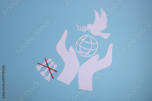 Paper hands on a blue paper background, a dove, a planet, and rockets crossed out with a red cross, all cut out of white paper. The idea is World Peace Day.