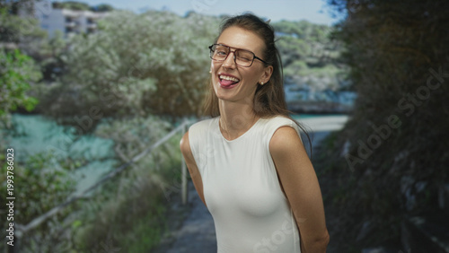Konstfotografi Woman with bare shoulders wearing glasses smiling and sticking tongue out on a shaded forest path by green water; playful joy