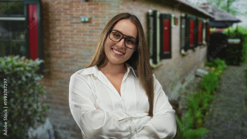 Obraz premium Woman wearing glasses smiling with arms crossed outside a brick building entrance; confidence welcome home.