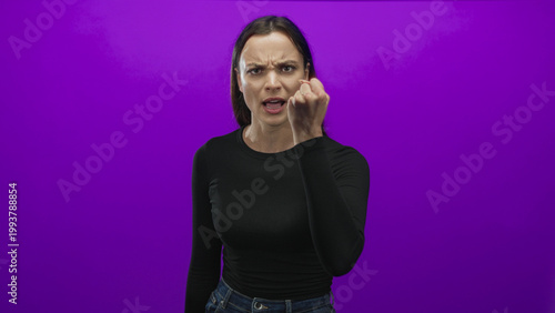 Фотография Woman in black long sleeve top and jeans beckons with curled hand and furrowed brow toward camera in studio with purple backdrop; anger defiance