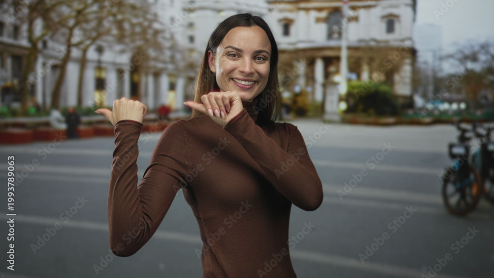Fototapeta premium Woman points thumb to a city street building while smiling and looking back, hands raised in a thumb gesture with visible nails; playful.