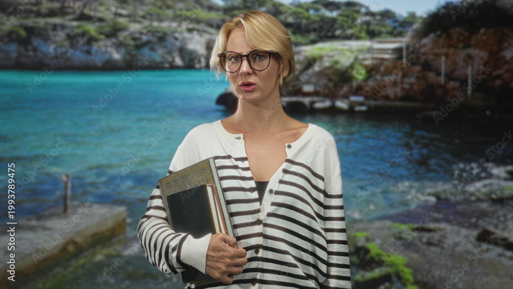 Fototapeta premium Woman holding book and notebooks, touching neck and adjusting glasses, striped sweater, posed before seascape backdrop in studio; thoughtful reflection.