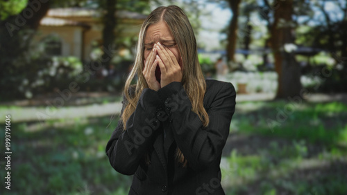 Fotografie Woman covering face with hands and rubbing her eyes while crying in front of a building and trees in an outdoor courtyard; grief loss