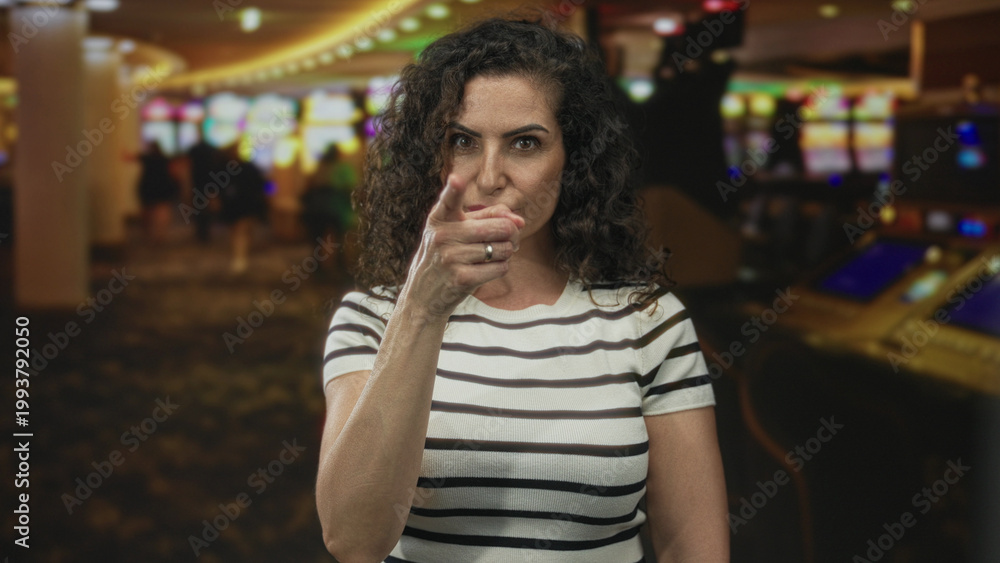 Fototapeta premium Young hispanic woman stares at camera and points finger at slot machine in casino building; determination.