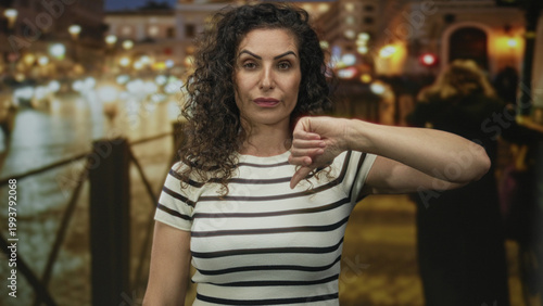 Fotografie Woman on a bustling city street holds out a hand with thumbs down gesture as traffic flows behind her; disappointment