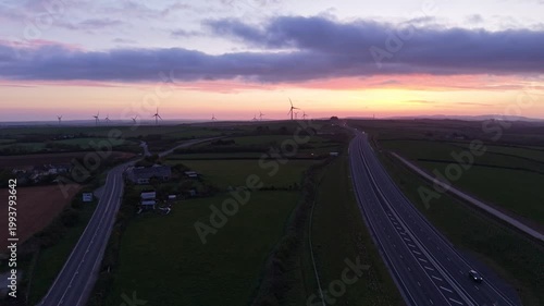 Aerial drone footage at sunrise showing the A30 dual carriageway cutting through the countryside, with wind turbines on the horizon beneath a vibrant, colourful sky