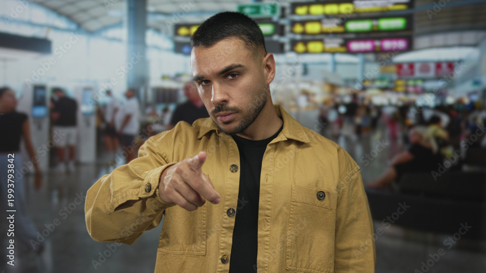 Naklejka premium Man with trimmed beard pointing finger directly at camera in crowded airport terminal with glass windows; confidence.