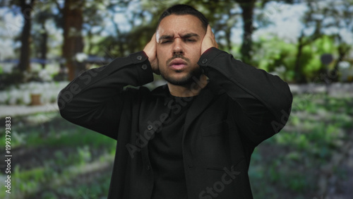 Canvas Print Young hispanic man covers his ears with both hands in sunlit forest clearing under tall green trees; stress