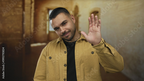 Canvastavla Hispanic man waving hand on urban street in daylight with casual posture and yellow jacket; welcome