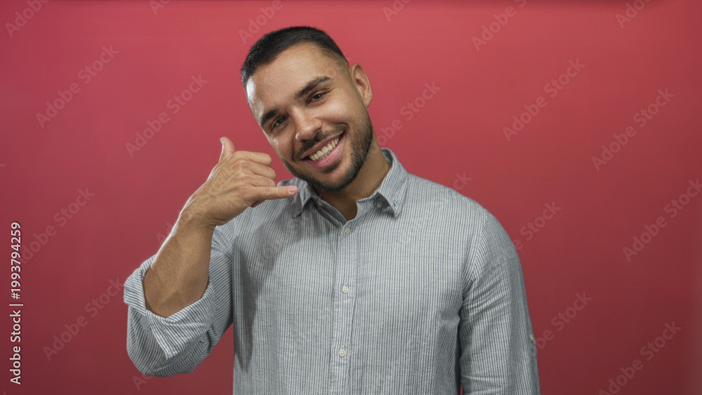 Fototapeta premium Man making phone gesture with his hand against red studio wall, smiling and winking as if calling someone; playfulness connection greeting communication.