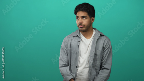 Quadro em tela Young hispanic man in casual shirt with hand on chest and hesitant expression in teal studio setting; apologetic regret