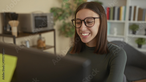 Fotografía Young woman adjusts eyeglasses and smiles toward a computer monitor while seated in a home studio with shelving, microwave and potted plant; concentration remote work