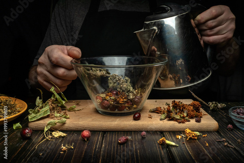 A person pours hot water from a kettle into a bowl filled with dry herbs and flowers. Various dried plants are scattered on the table around them. The work takes place in a dark kitchen