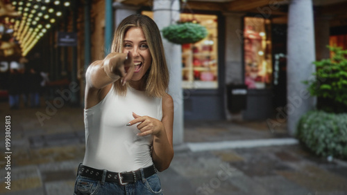 Obraz na płótnie Woman points finger at camera on a street storefront with string lights, wearing white sleeveless top and jeans; playful