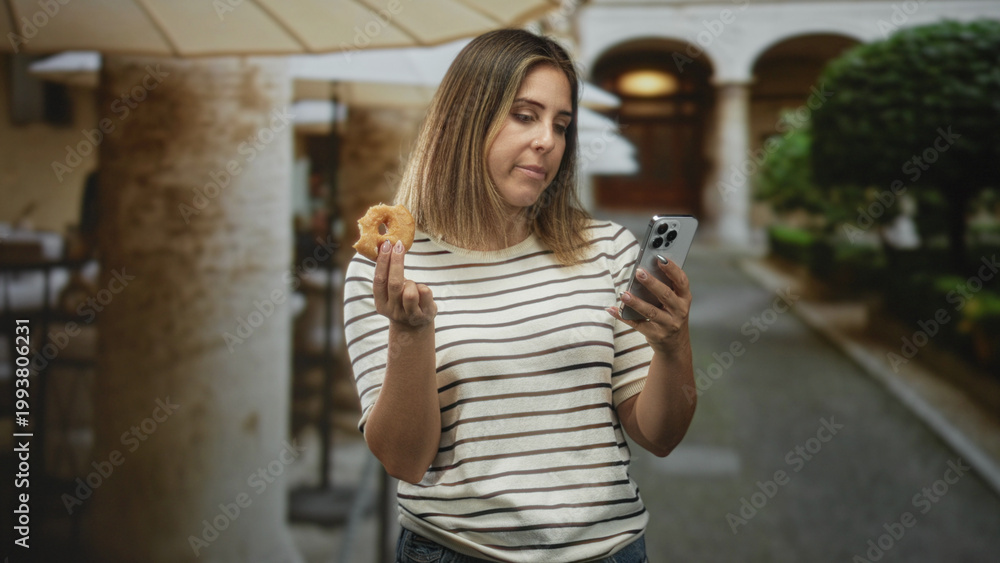 Fototapeta premium Woman holding a donut in her hand and checking a smartphone while standing under a colonnade by a street; distraction.