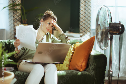 An overwhelmed woman sits on a velvet couch, hand on forehead, grappling with papers and a laptop. The scene powerfully depicts the mental strain of financial worries or intense remote work from home.