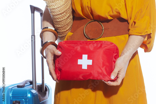 A woman in a vibrant yellow dress prepares for her summer vacation by packing a red medical first aid kit next to her blue hardshell suitcase, prioritizing travel health and safety.