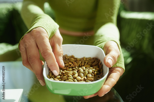 A woman in a green top mindfully selects pumpkin seeds from a small bowl, emphasizing the importance of healthy ingredients in a wellness routine.