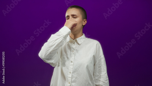 Foto Woman pinches nose with hand wearing white shirt in studio purple backdrop; disgust smell reaction unpleasant