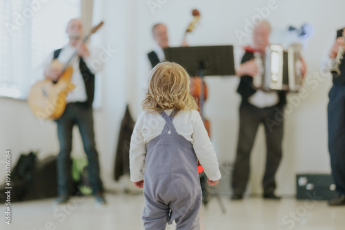 Toddler stands captivated while watching a live band perform indoors. Musicians play guitar, accordion and bass in soft focus background, creating a warm family-friendly concert atmosphere.