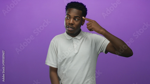 Billede på lærred Young african american man in white polo shirt points finger to temple in studio against purple studio wall; playful madness