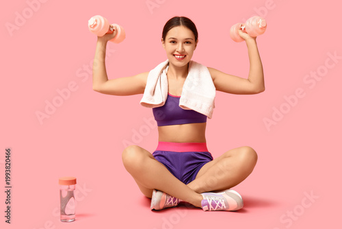 Young woman in sportswear and with dumbbells sitting against pink background