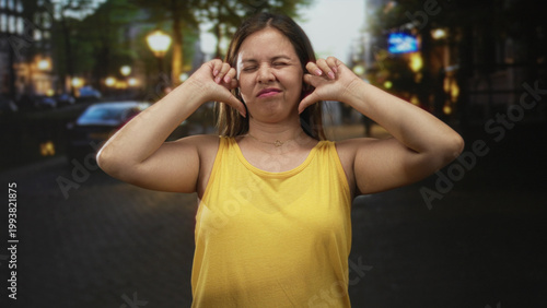 Bild auf Leinwand Woman plugs ears with fingers and eyes closed on busy city street at dusk wearing yellow tank top and necklace; annoyance urban noise