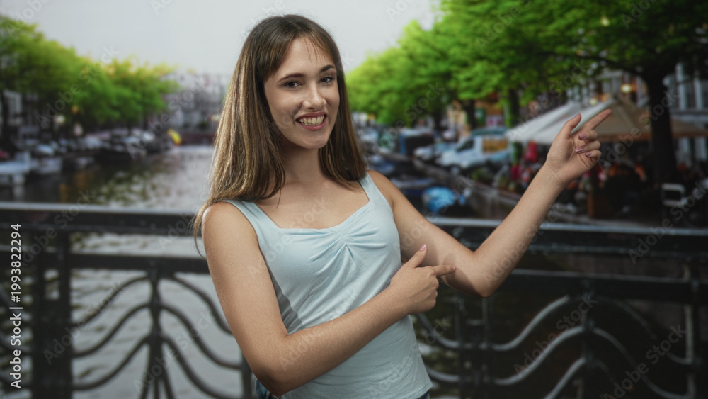 Fototapeta premium Woman in light blue tank top points finger at canal cafe from a street bridge in amsterdam with trees and outdoor seating; joy.