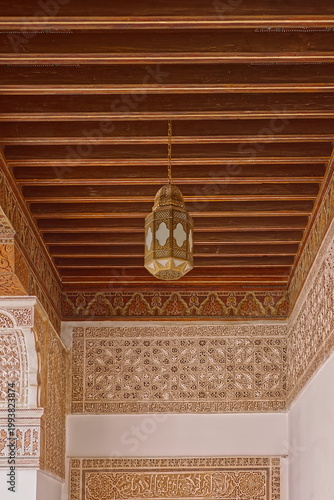 Brass lamp pending from the painted wood ceiling of the carved stucco-frieze topped NW gallery, Ben Youssef Madrasa courtyard. Marrakesh-Morocco-315