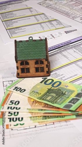 Close-up of euro banknotes laid on construction blueprints and cost sheets while a realtor places house models one by one, symbolizing real estate development, budgeting, and investment planning. 