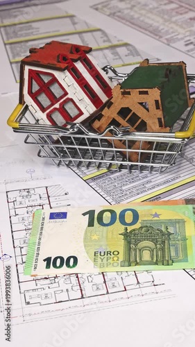 Close-up of house models placed in a grocery shopping basket while euro cash is being counted over apartment floor plans, symbolizing real estate buying decisions, budgeting, and investment planning. 