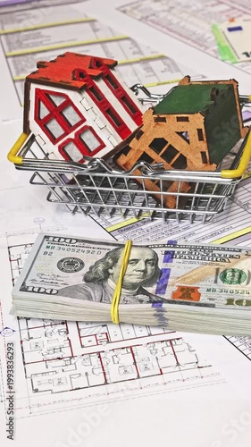 Close-up of house models in a shopping basket while a stack of US dollars is placed and keys are added on top, symbolizing real estate purchase, ownership transfer, and investment deal closure. 