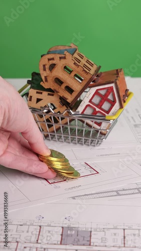Vertical footage showing a hand placing bitcoin coins on architectural blueprints next to a shopping basket holding three miniature houses, filmed on chroma key. Represents real estate investment