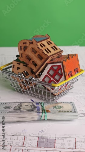Vertical footage of a stack of US dollar bills placed in front of a shopping basket holding miniature house models on architectural blueprints, filmed on chroma key. Concept of real estate investment