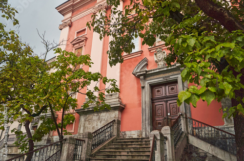 Entrance to the Franciscan Church of the Annunciation. The facade of a pink church with green trees on the sides in Ljubljana, Slovenia. High quality photo
