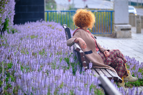 Stylish redhead woman surrounded by purple flowers in urban park setting
