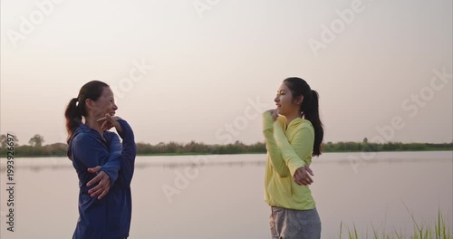 Happy Asian mother and teenage daughter stretching by lake, mindful movement in evening golden light, authentic family wellness
