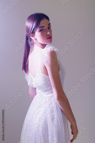 Young woman in a white lace dress poses with her back turned, looking over her shoulder. Soft lighting highlights her beauty and fashion style in a studio setting.