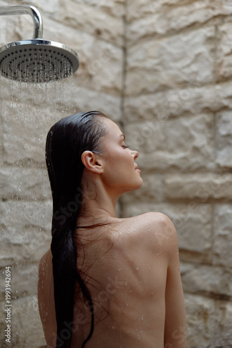 Woman showering with eyes closed under rainfall showerhead, wet hair cascading down back, serene expression, natural stone wall background, wellness moment.