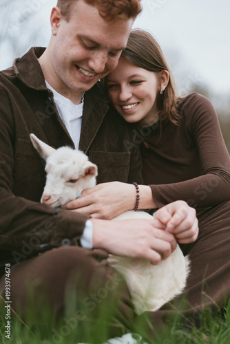 Couple outdoors, smiling together while cuddling a small goat kid in a sunlit meadow, warm lighting, casual clothing, gentle breeze, natural colors, rustic background, candid lifestyle moment
