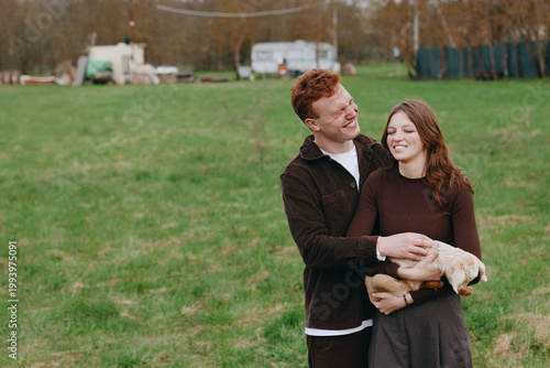 Happy couple outdoors in a grassy field, embracing with a small dog between them, warm affectionate moment, casual clothing, candid lifestyle scene, serene rural backdrop