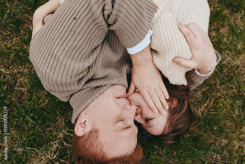 Tender moment between parent and child lying on grass, faces close, hands touching softly, outdoor lifestyle, warm sunlight, gentle affection, candid connection in a peaceful park setting.