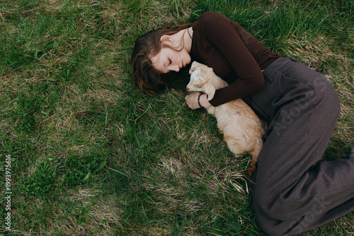 A calm lifestyle scene shows a woman lying on grass, cuddling a gentle goat kid, expressing warmth, tenderness, and connection with nature in an outdoor setting, conveying peaceful companionship and