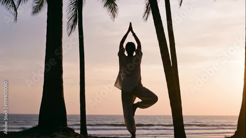 Panning slow motion shot of person practicing tree pose yoga on palm lined tropical beach at sunset overlooking calm ocean